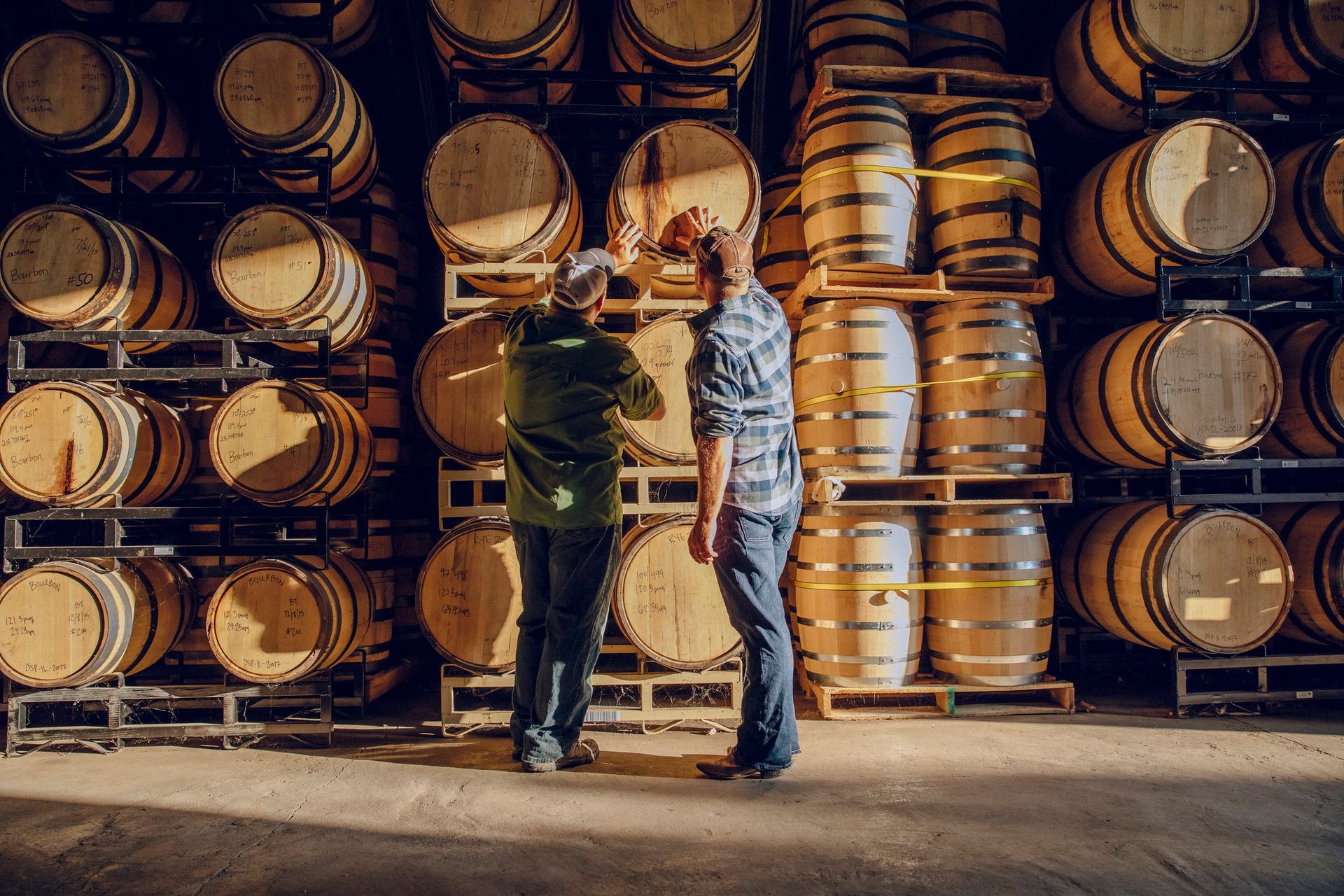 Two people inspecting wooden barrels in a distillery warehouse.