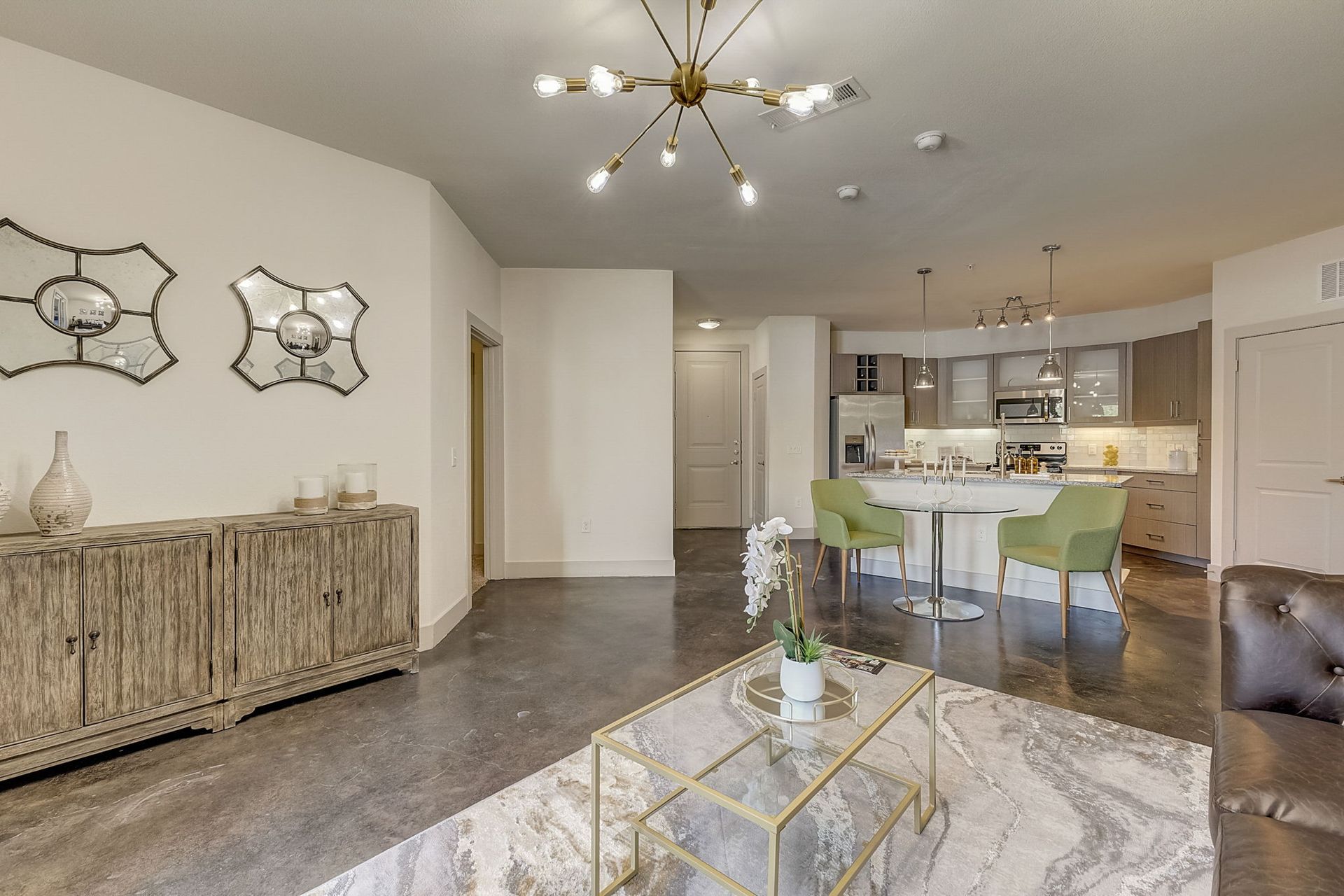 Living room with a concrete floor, open to a kitchen with a breakfast bar. A wooden cabinet, mirrors, and a gold coffee table are present.