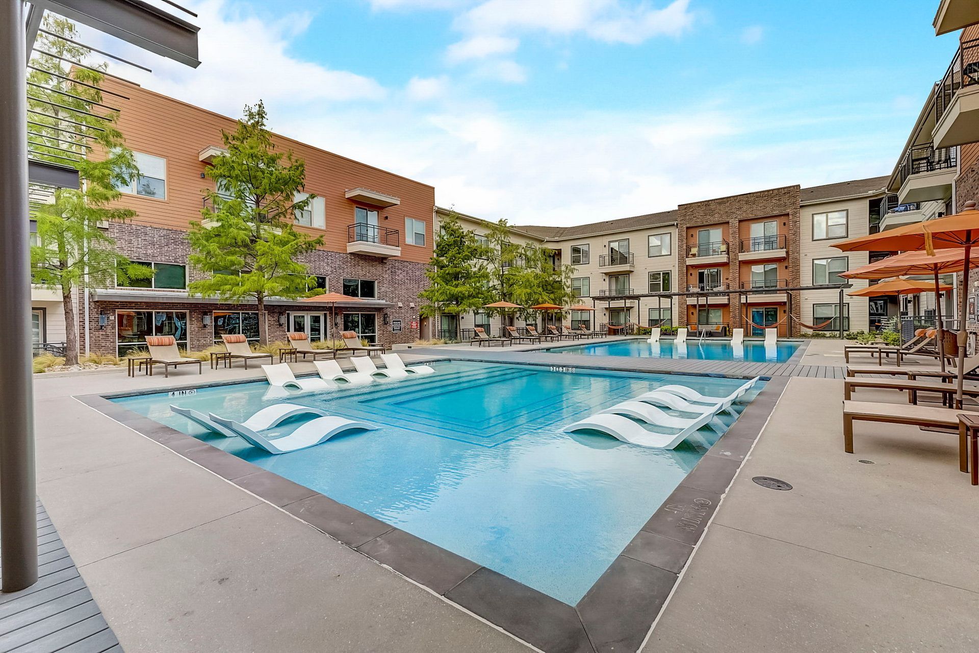 Swimming pool surrounded by apartment buildings under a blue sky.  Chairs and umbrellas are poolside.