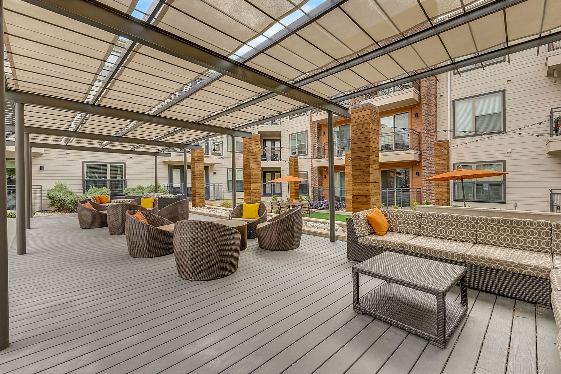 Outdoor seating area with brown wicker furniture under a beige pergola. The patio is wood-paneled, with buildings visible in the background.