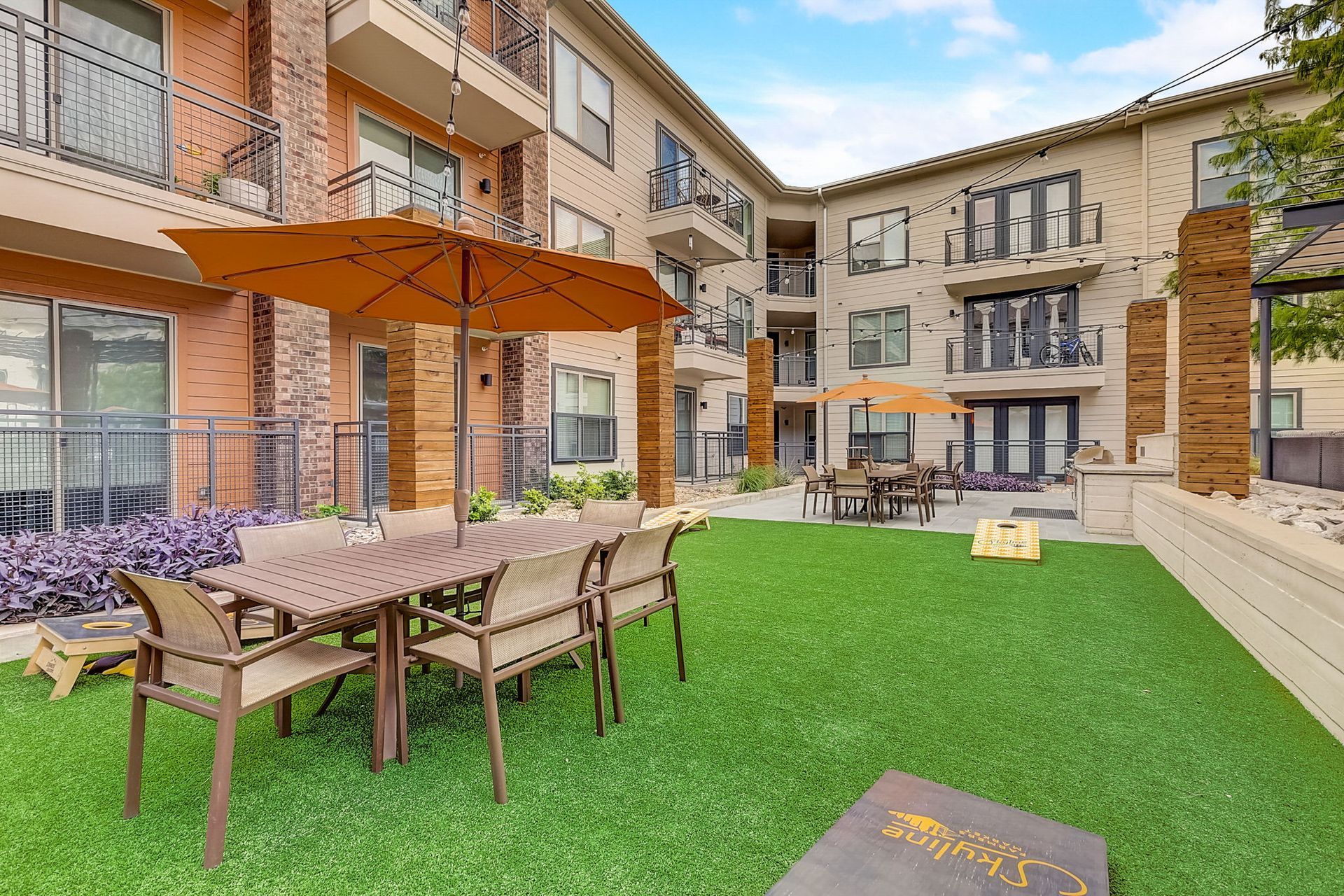 Outdoor patio area with tables, chairs, umbrellas, and artificial grass in front of a multi-story building.
