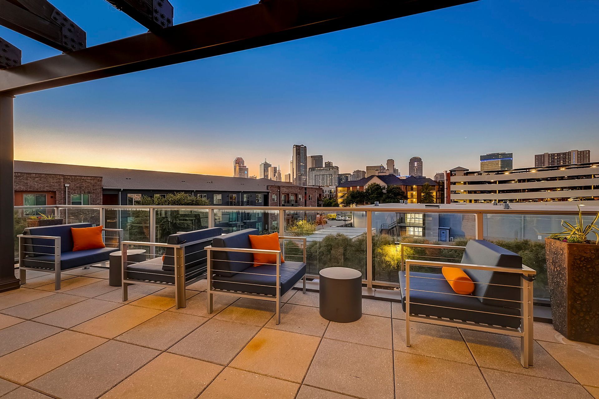 Rooftop patio with modern furniture overlooking a cityscape at dusk; blue couches with orange pillows, gray stone patio, and glass railing.