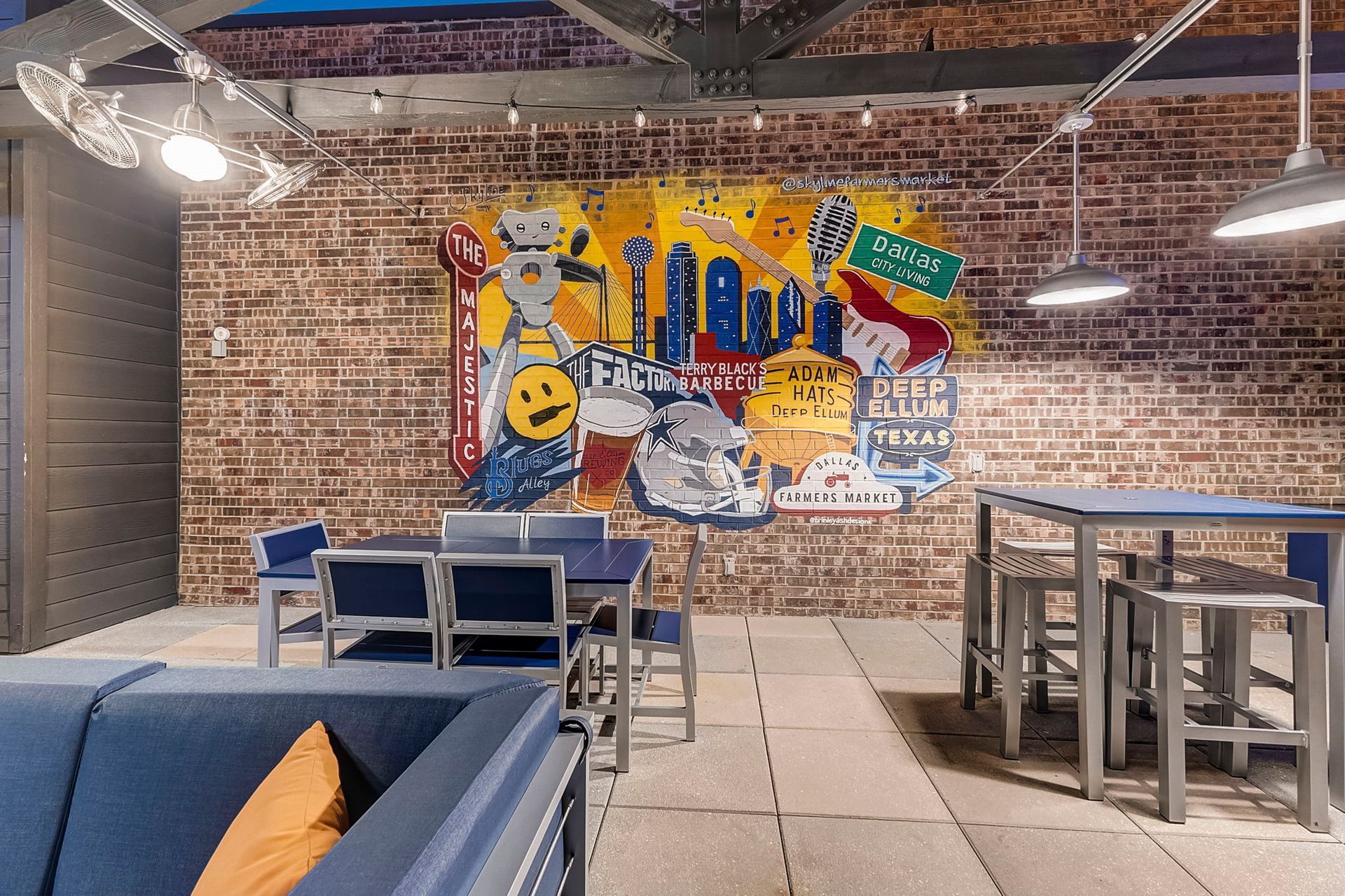 A modern interior with brick walls, a colorful mural, and seating areas with blue furniture. Overhead lighting.