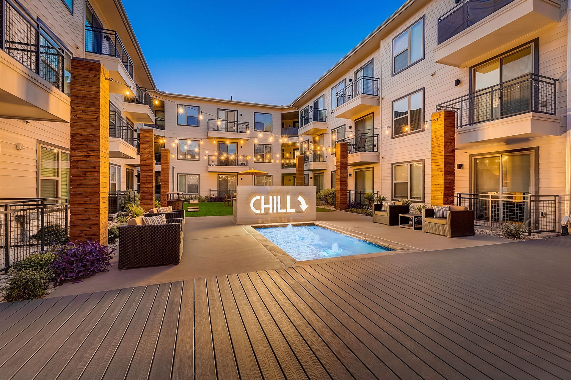 Courtyard of an apartment complex with a reflecting pool, seating areas, and string lights under a blue sky.
