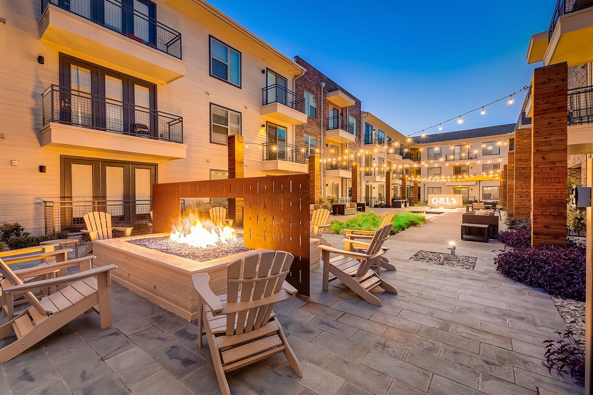 An outdoor patio with a fire pit and lounge chairs in front of a multi-story building at dusk.