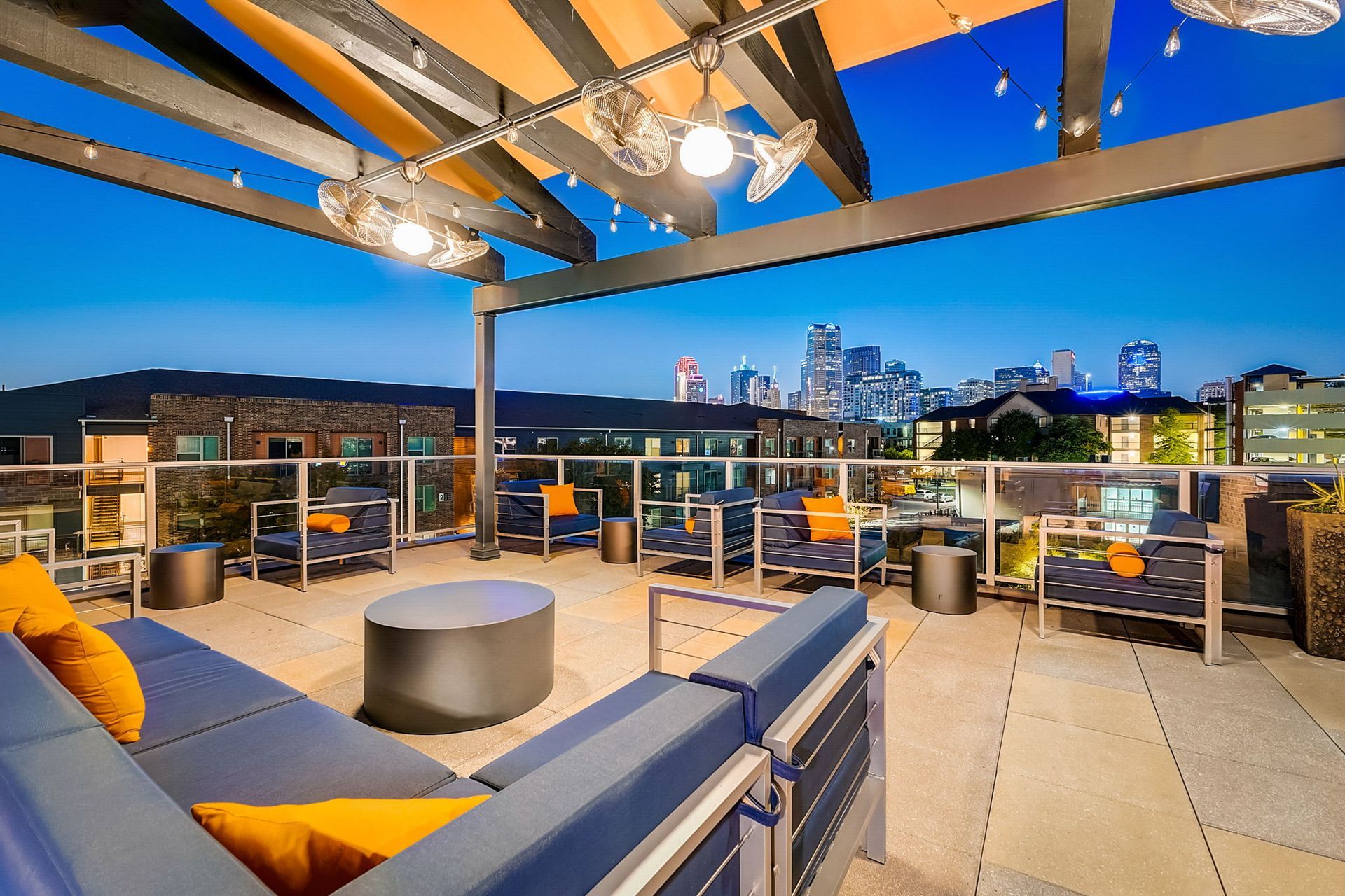 Rooftop patio with lounge furniture overlooking a city skyline at dusk. Blue and orange accents, string lights.