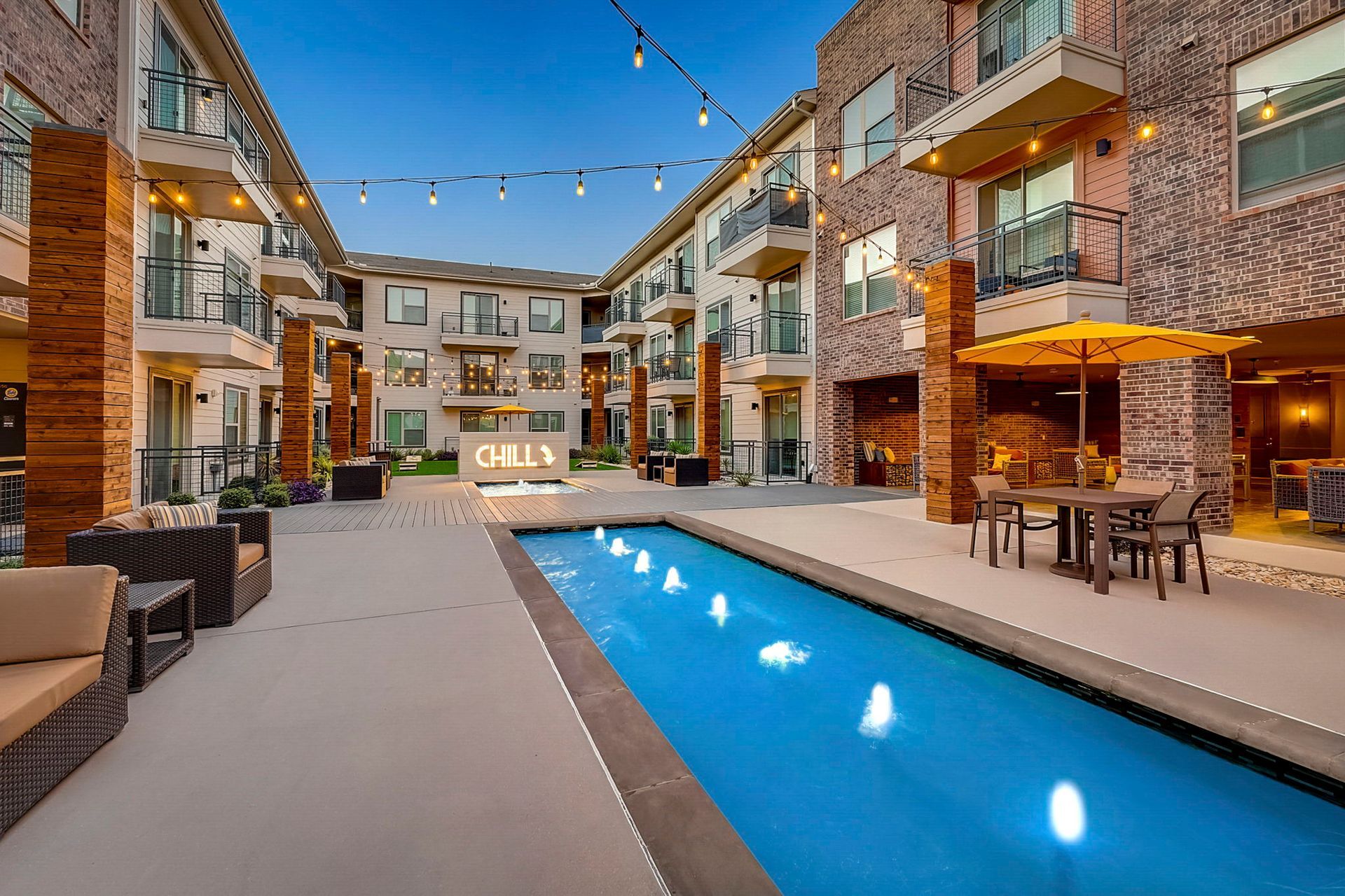 Courtyard of a modern apartment complex with a pool, seating, and brick facade. String lights hang overhead.