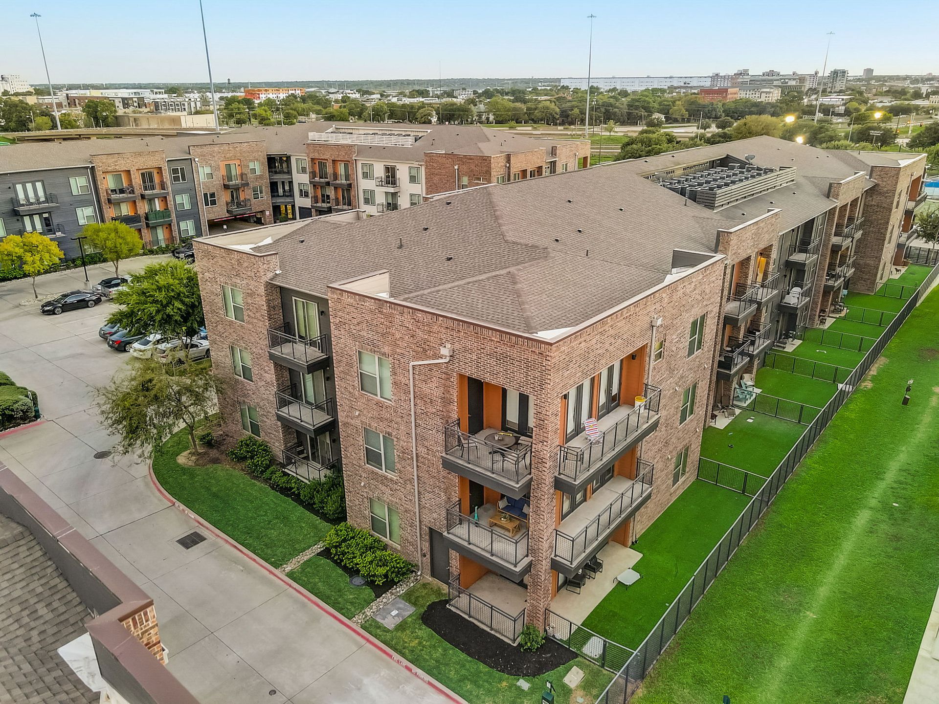 An aerial view of a brick apartment building with balconies, surrounded by green grass and a parking lot.
