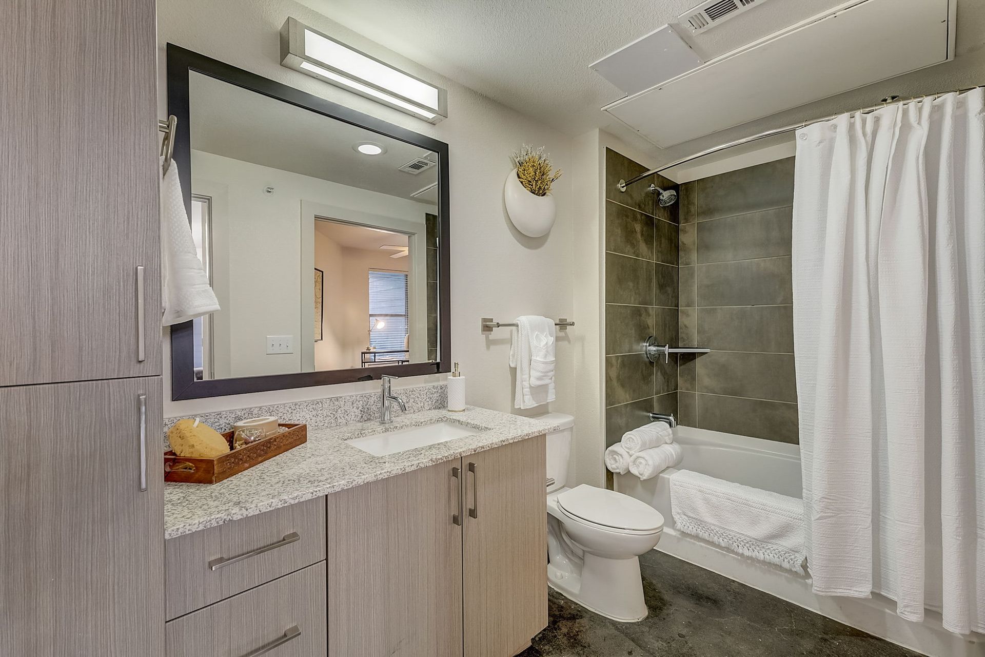 Modern bathroom with a granite countertop vanity, large mirror, and shower/tub combo. White shower curtain, and light grey cabinets.