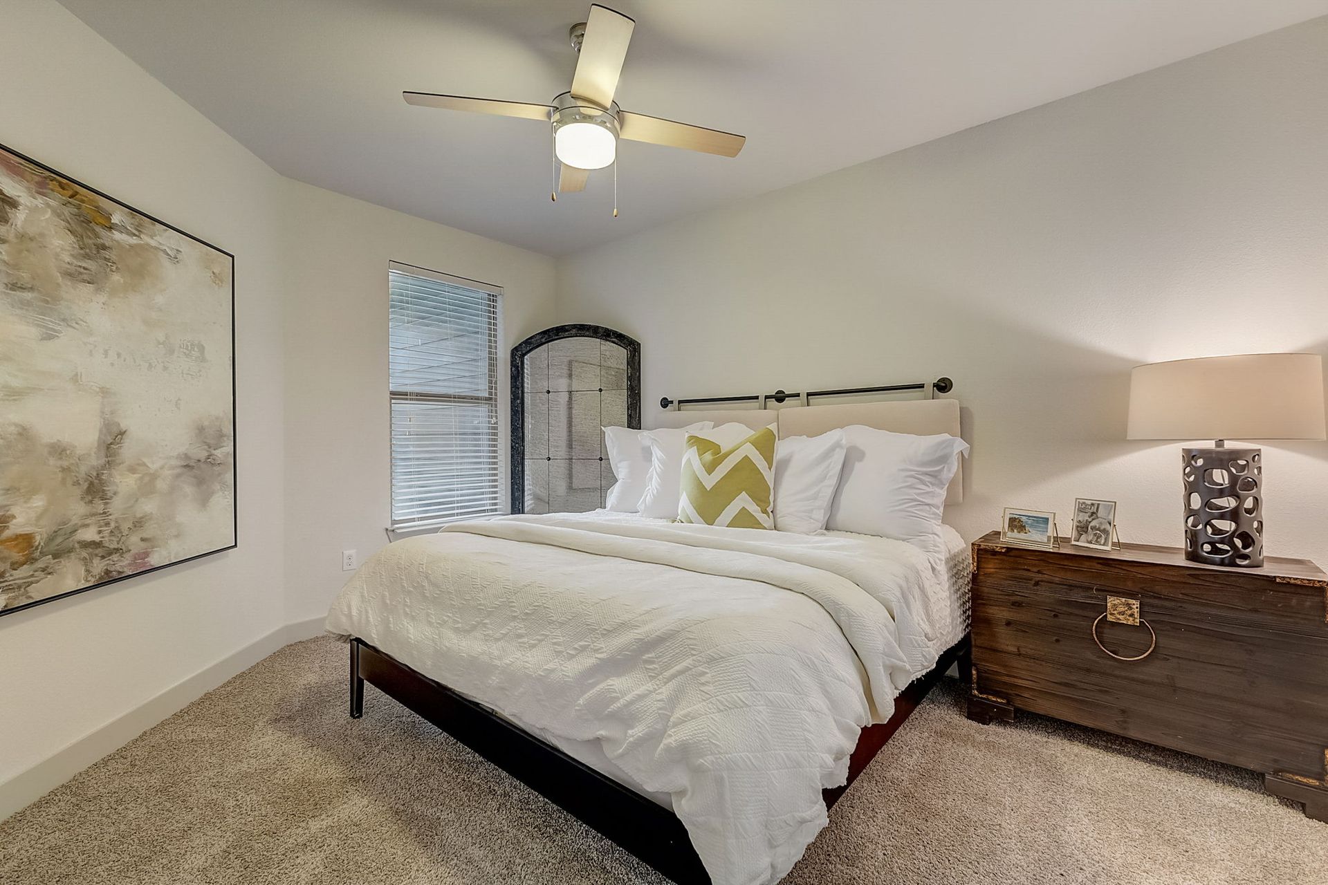 Bedroom with a bed, artwork, a trunk-like nightstand, and a decorative mirror; neutral tones.