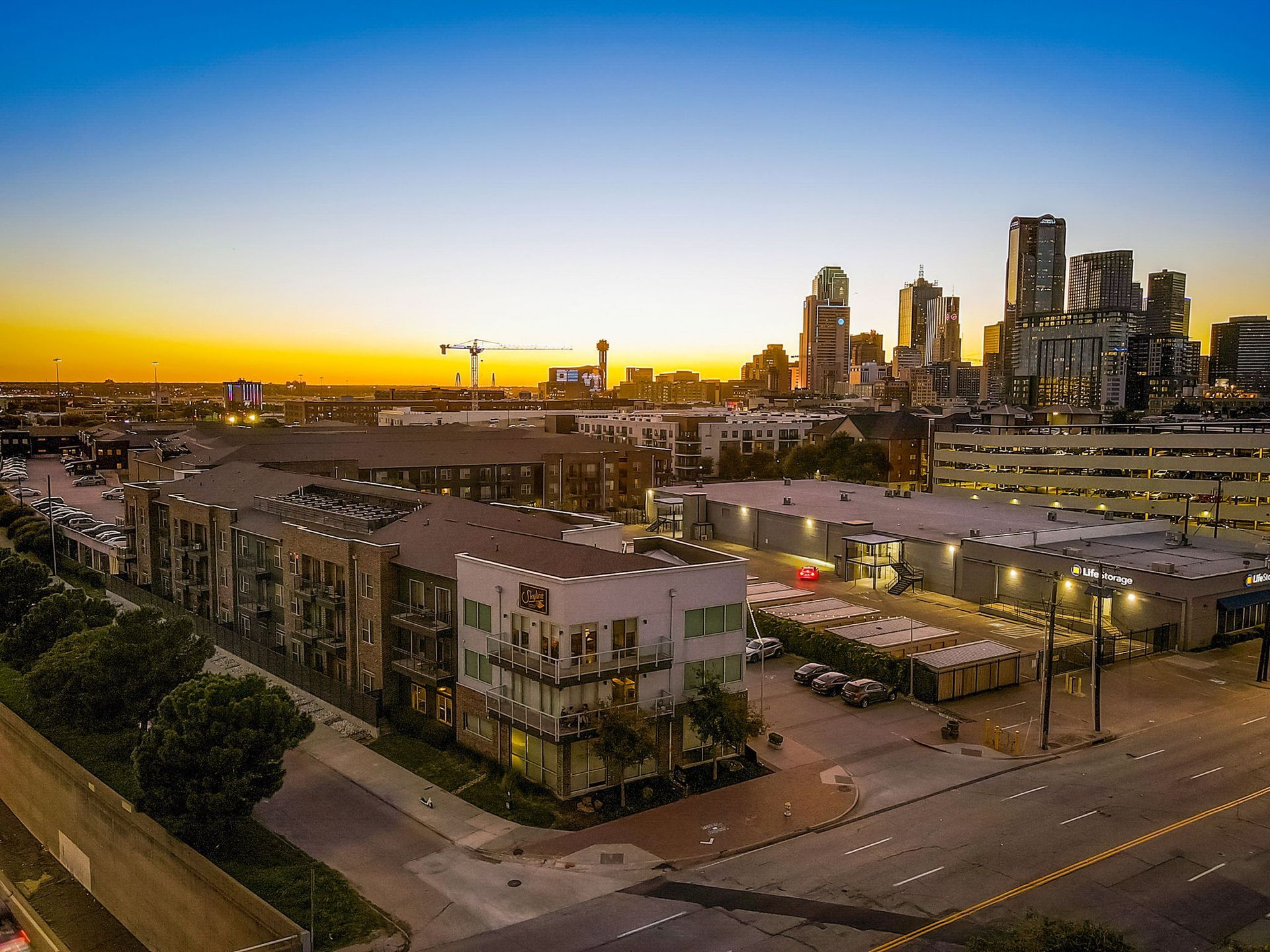 Aerial view of Dallas skyline at dusk, buildings in foreground, with colorful sunset.
