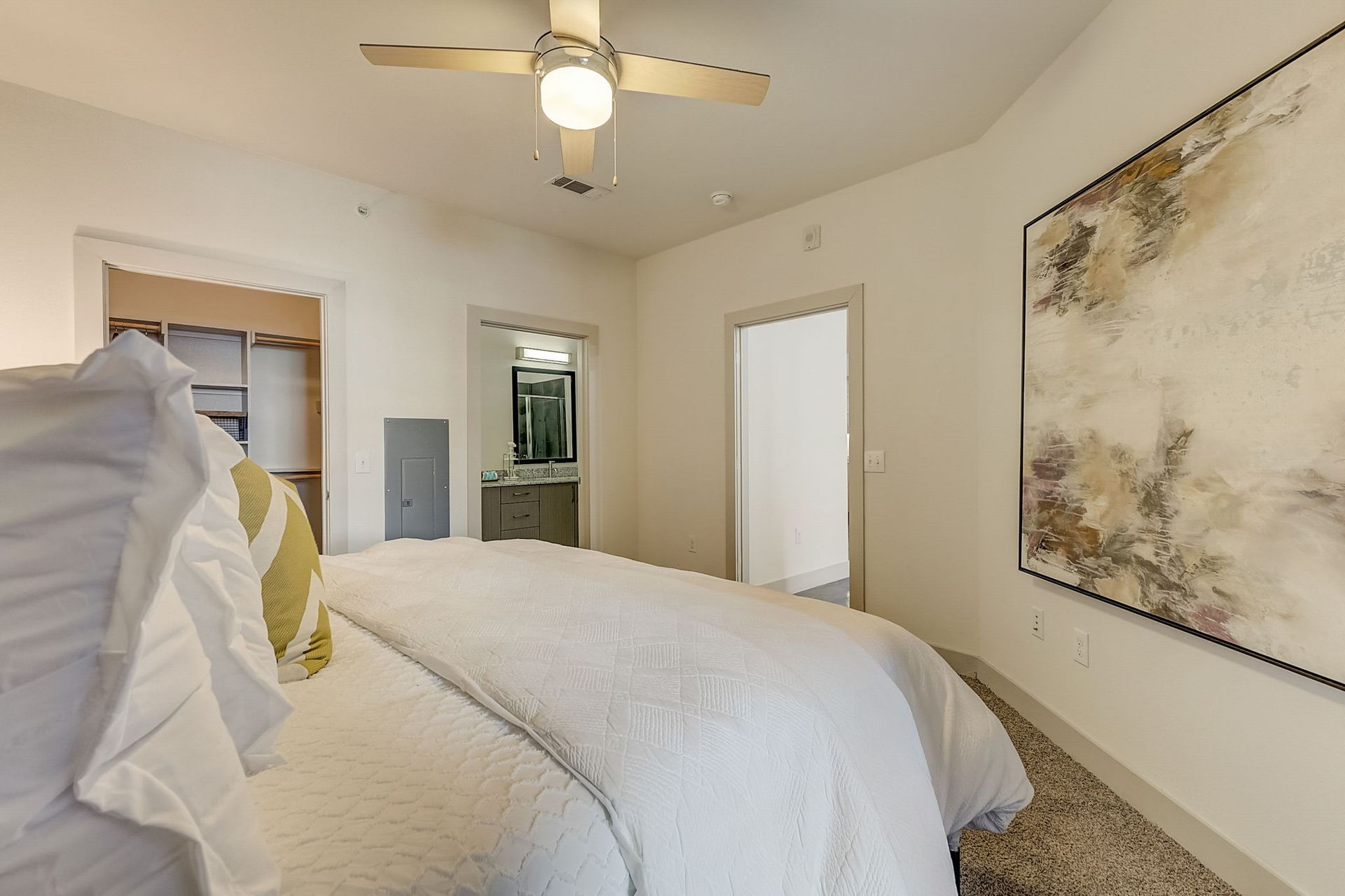 Bedroom with a white bed, art on the wall, and doors leading to a walk-in closet and bathroom. A ceiling fan hangs overhead.
