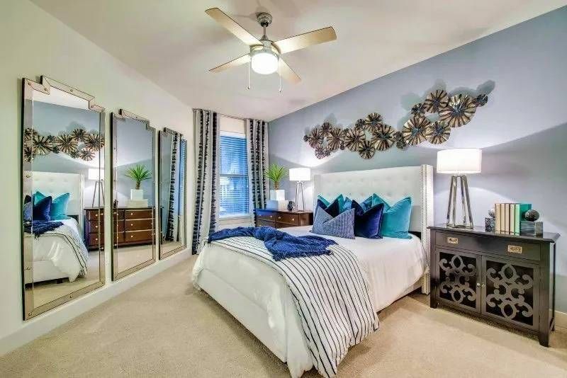 Bedroom with blue and beige decor. Features include a bed, pillows, side tables, large mirrors, and a metal wall art piece.