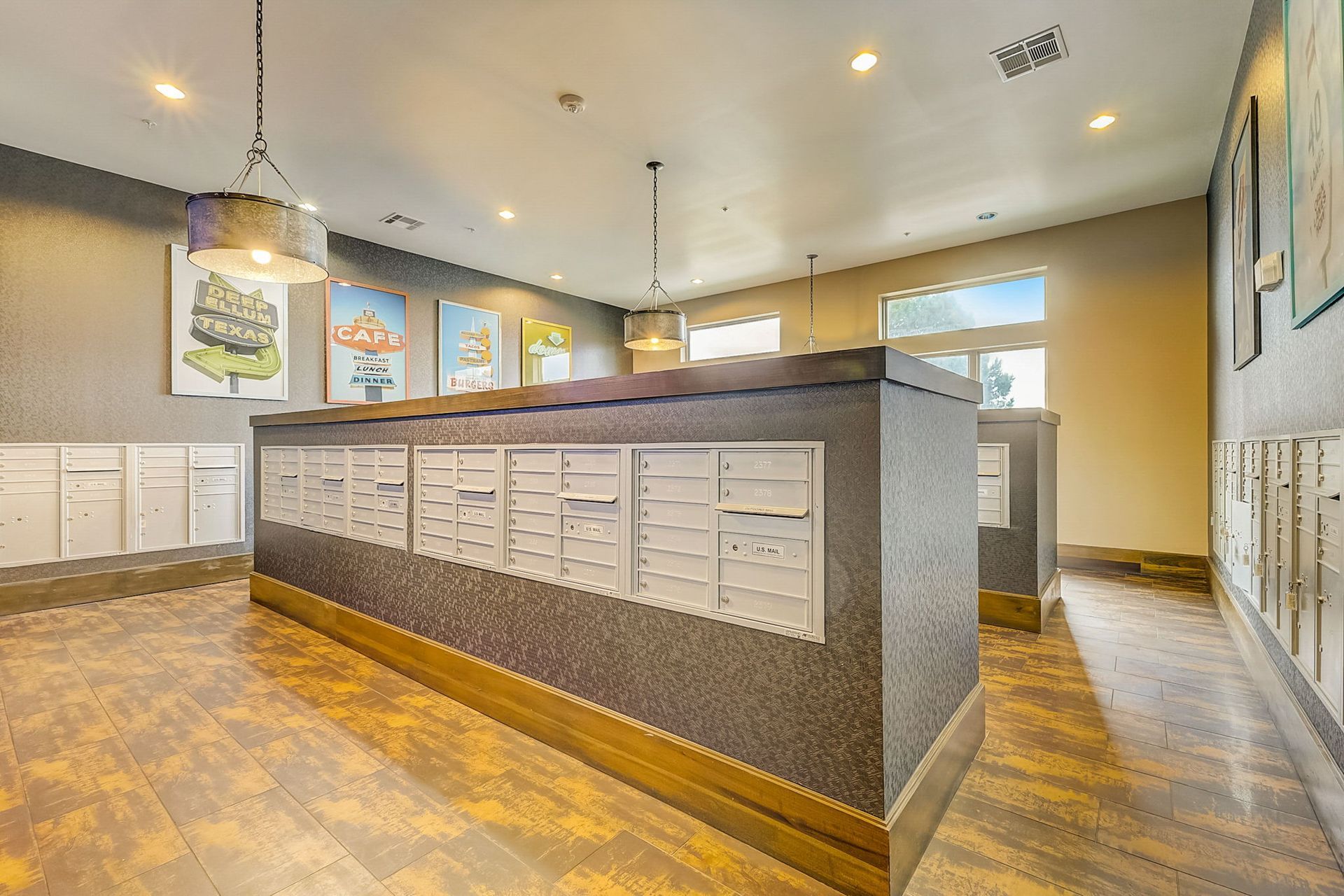 Apartment mailroom with rows of mailboxes, gray walls, wood flooring, and artwork. Three pendant lights hang from the ceiling.