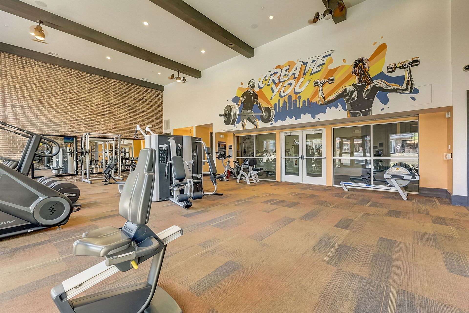 Gym interior with various exercise machines, a colorful mural, and large windows.  Orange and brown tones dominate the space.