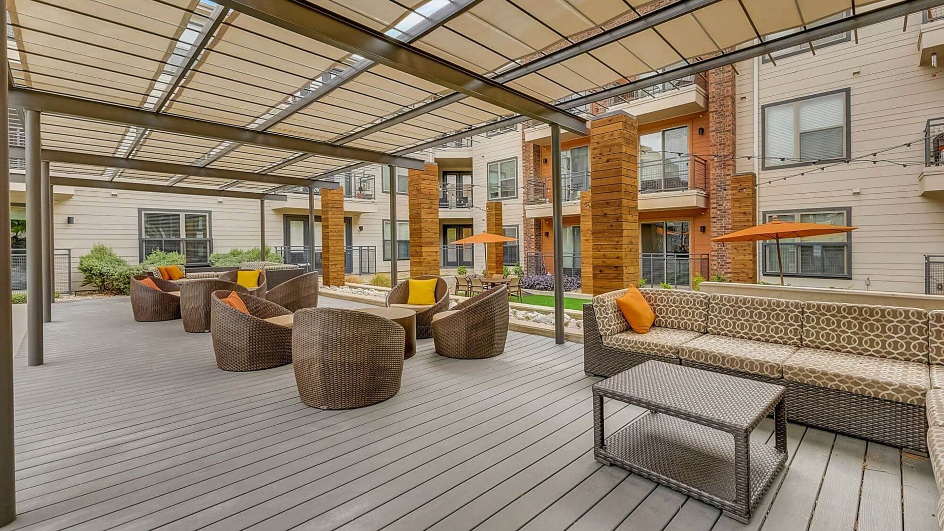 An outdoor patio with seating and a pergola. Neutral-toned wood decking, wicker furniture, and brick columns under a canopy.
