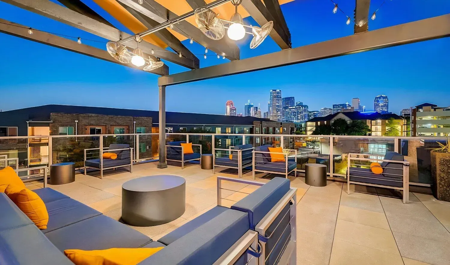 Rooftop lounge with city skyline view at dusk. Blue and orange seating, string lights, and a modern, open-air design.