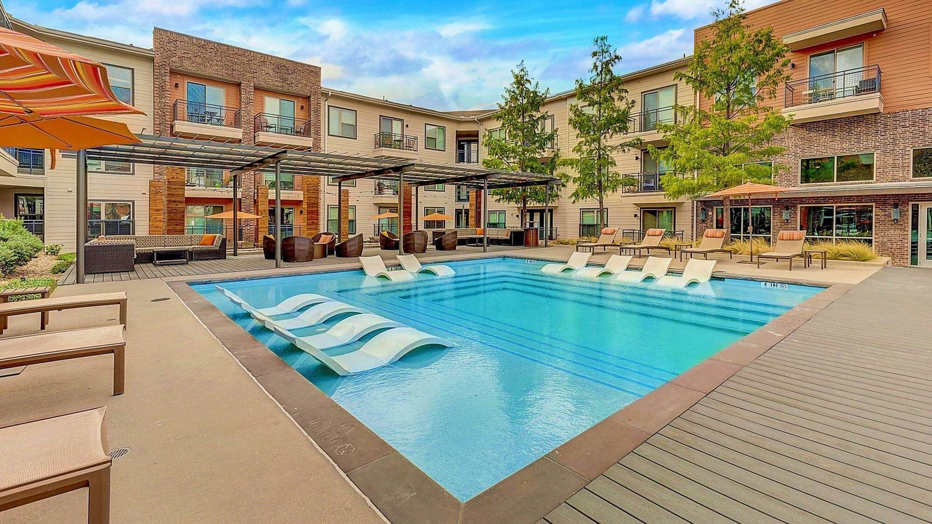 Swimming pool in a courtyard surrounded by apartment buildings with lounge chairs and pergolas. Blue sky overhead.