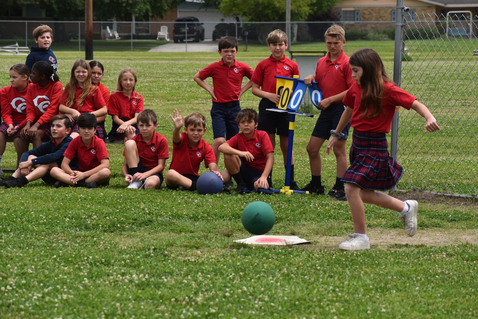 A group of children in red shirts are sitting on the grass watching a girl throw a ball