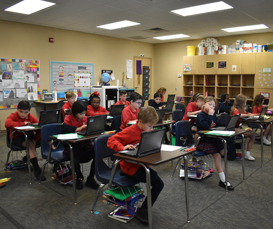 A group of children are sitting at desks in a classroom using laptops