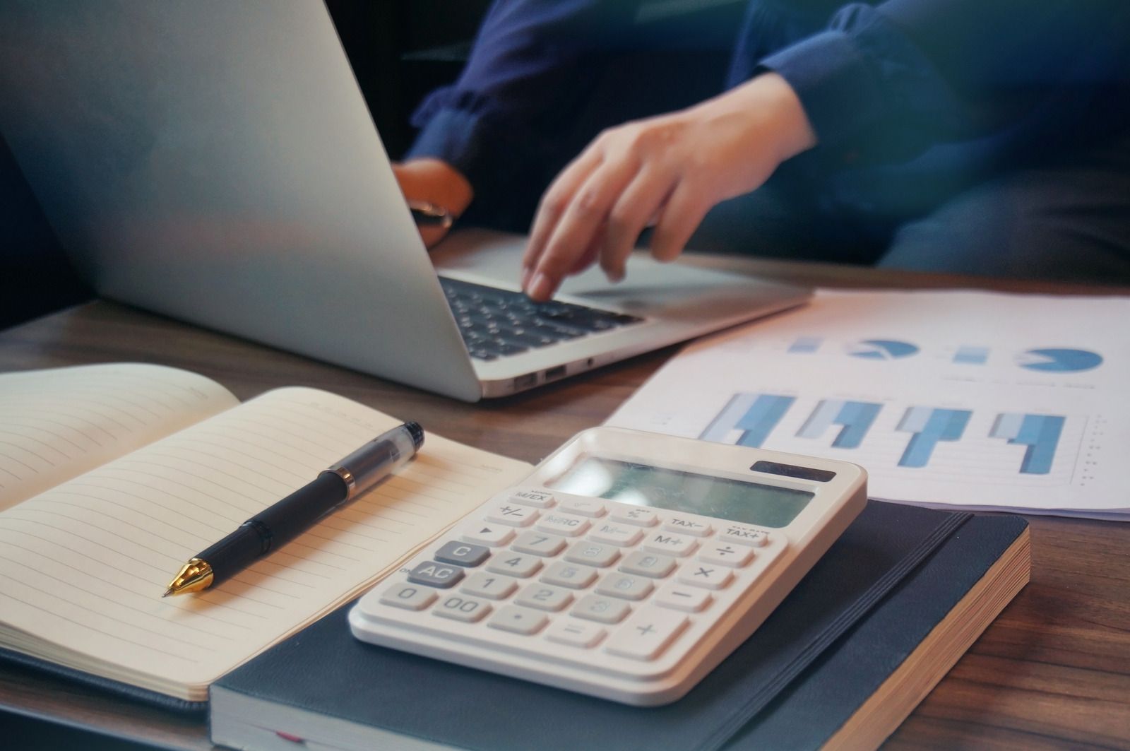 Person typing on laptop, working with a calculator, notepad, and graphs on a desk.