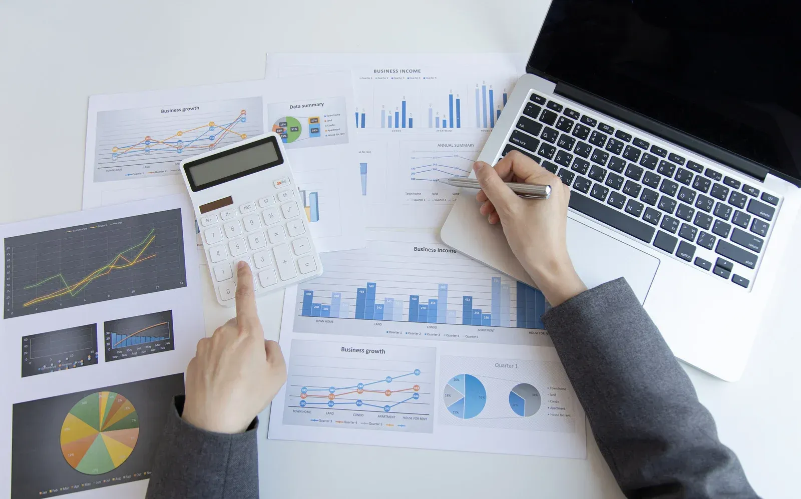Person working on financial documents with a calculator and laptop. Blue and yellow charts, hands working.