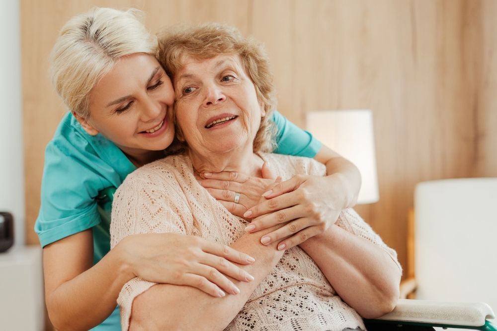 Woman in teal uniform hugs an elderly woman seated in a wheelchair; they both smile.