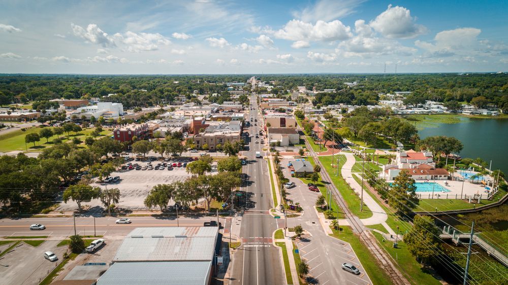Aerial view of a downtown street lined with buildings, green trees, and a lake under a cloudy sky.
