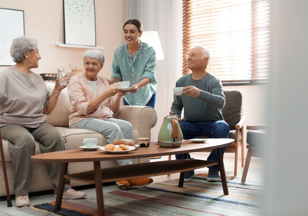 A caregiver serves tea to a group of seniors sitting in a living room.