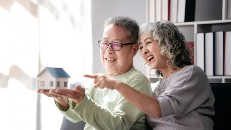 Senior couple holding house model for buying in life retirement while sitting on the couch and spending time to doing activity for relaxation together at living room of home.
