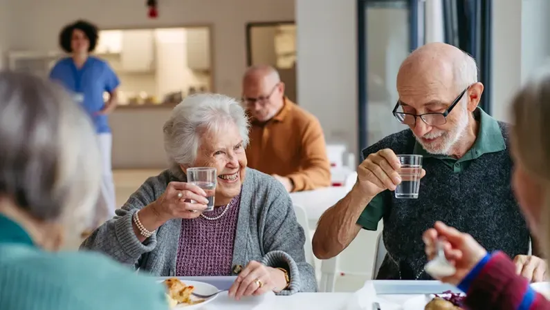 Group of seniors smiling and drinking water at a dining table in a care facility.