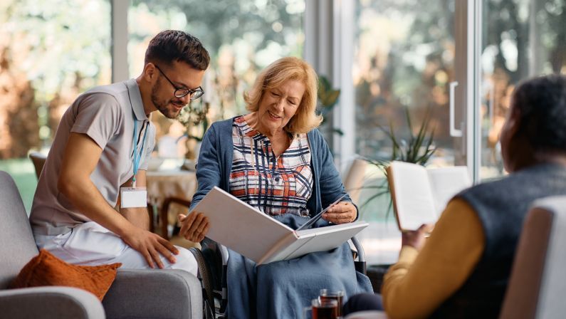 Happy elderly woman in wheelchair looking at her family photos in album with young caregiver in residential care home.