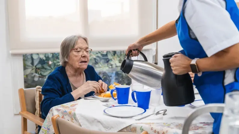 Caregiver pouring hot beverage for an elderly woman during breakfast at a nursing home, highlighting daily care and support in a geriatric setting