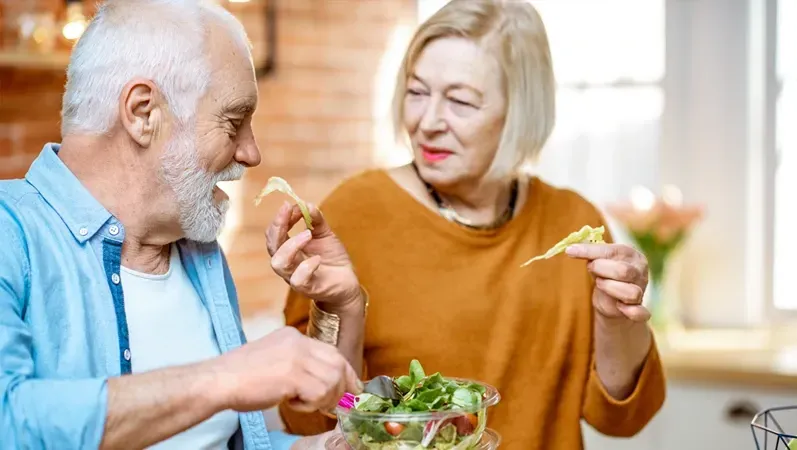 Elderly couple eating salad, smiling, in kitchen. Woman wears orange, man in blue shirt.