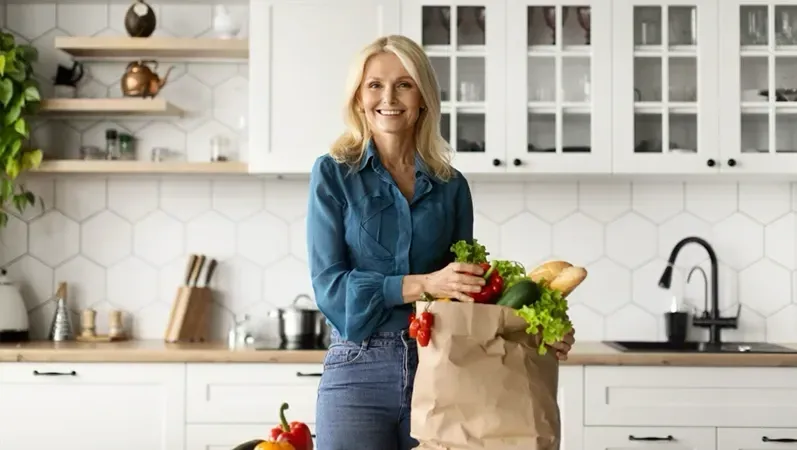 Woman in a kitchen holding a paper bag of groceries, smiling.