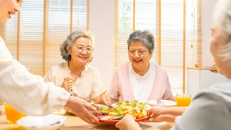 Woman serving a plate of food to smiling seniors at a dining table.