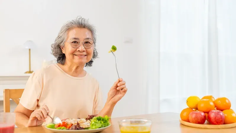 Elderly couple eating salad, smiling, in kitchen. Woman wears orange, man in blue shirt.