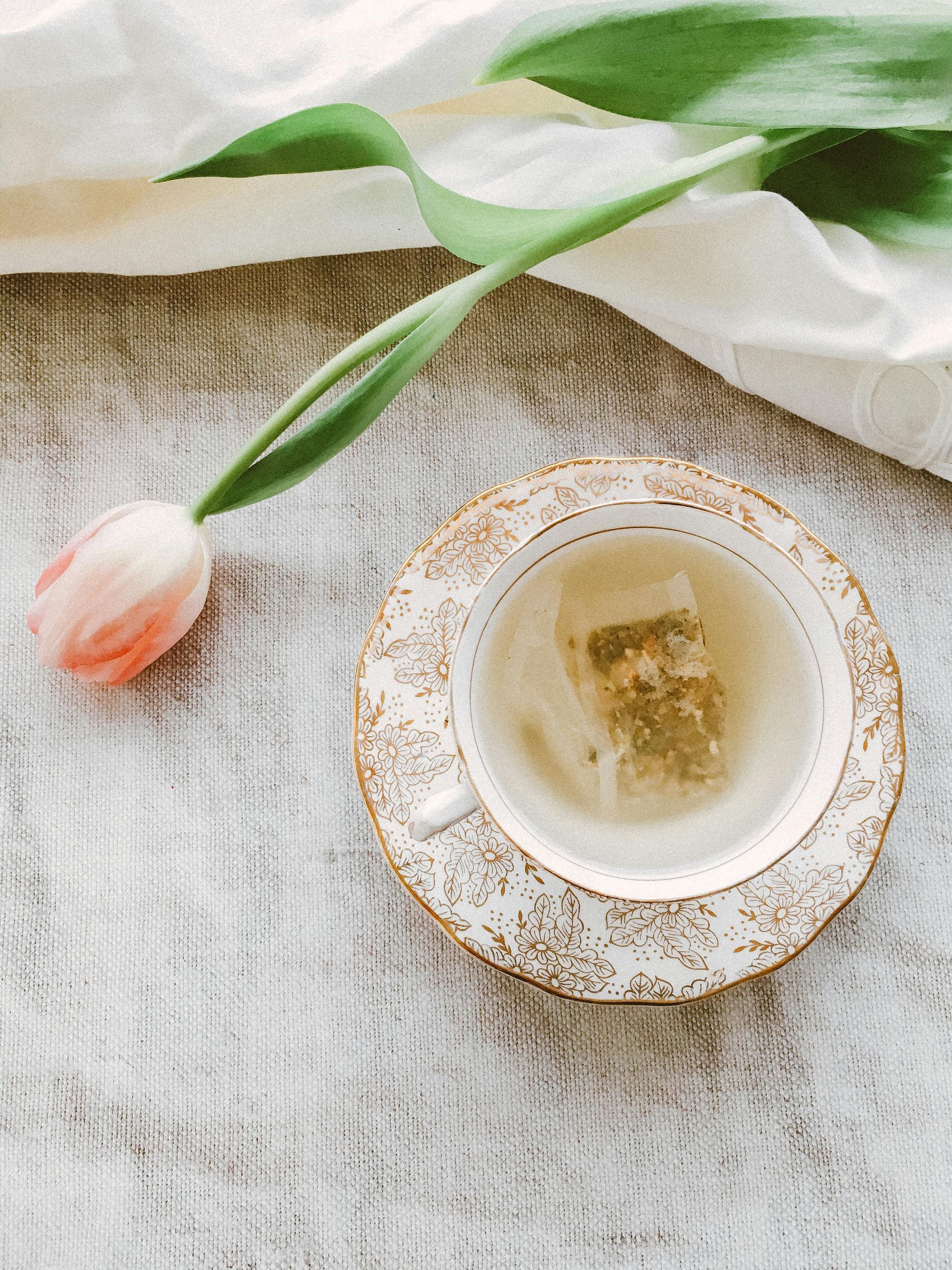 A teacup with tea and tea bag on a decorative saucer, tulip on a white textured surface.