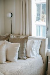 Bed with various white and tan pillows, next to a window with light curtains.