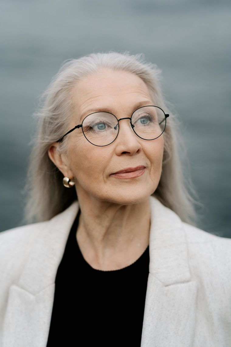 Senior woman with gray hair wearing glasses and a white blazer, looking up.