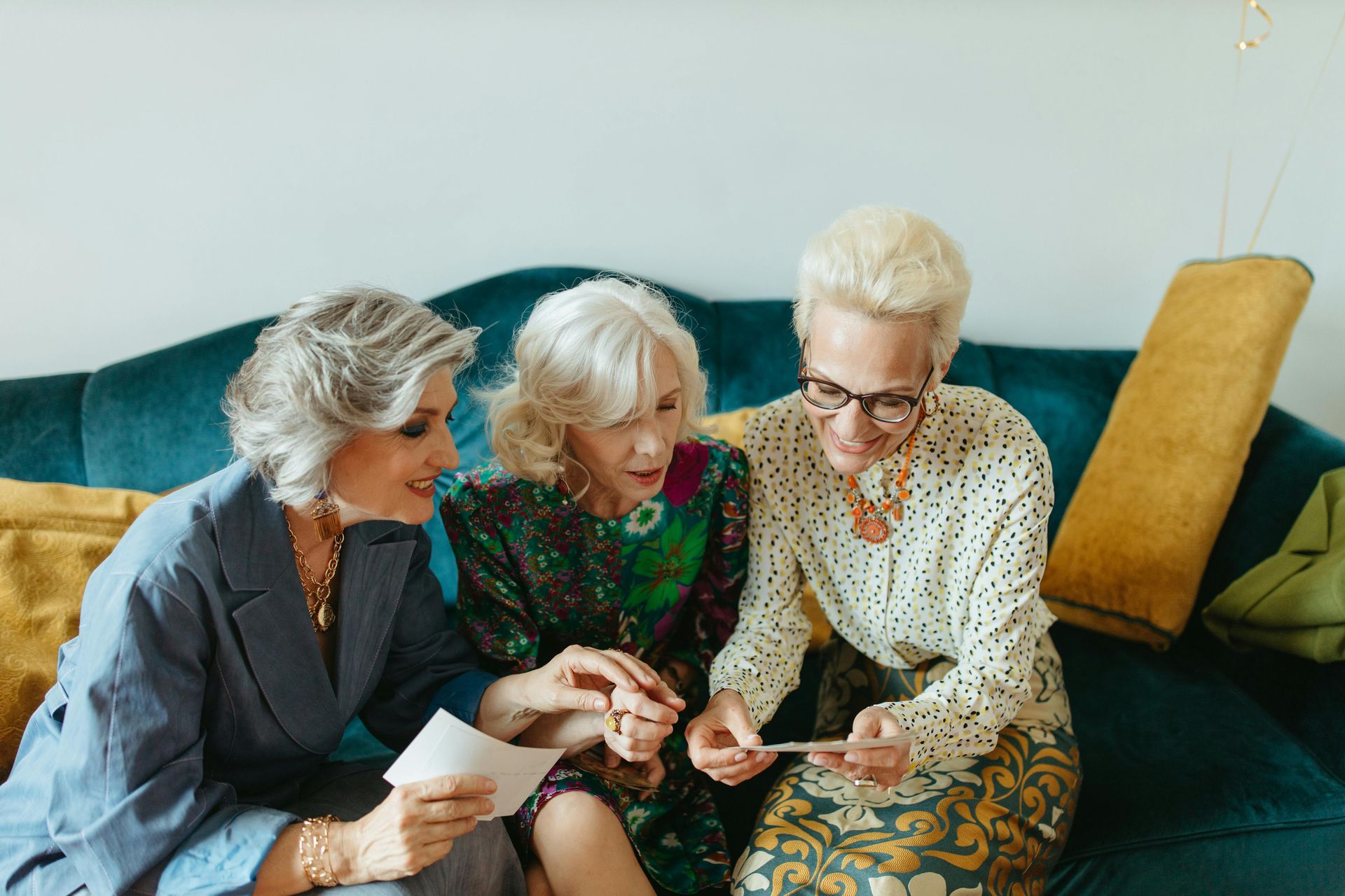 Three older women on a blue sofa look at photos. One holds pictures; all appear engaged and smiling.