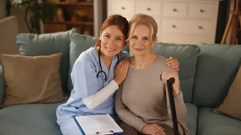 A home healthcare nurse in scrubs smiles while embracing a senior woman who is holding a cane. They are sitting on a sofa in the woman's living room during a daytime visit.