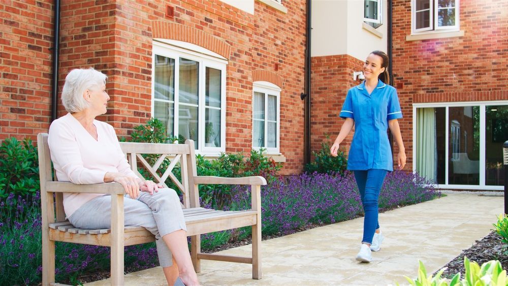 Senior Woman Sitting On Bench And Talking With Nurse In Retirement Home