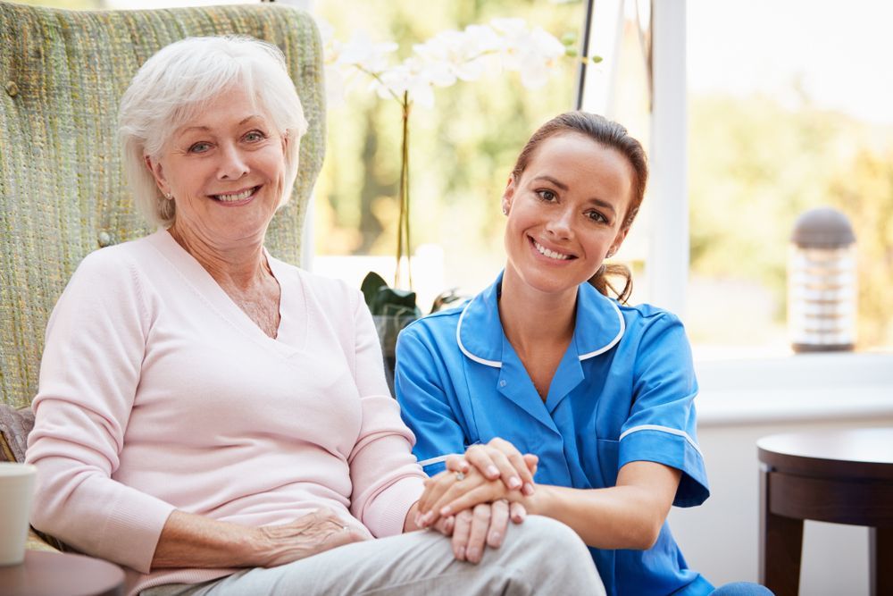 Senior woman and healthcare worker smiling together indoors. The woman is seated, the worker's hand on hers.