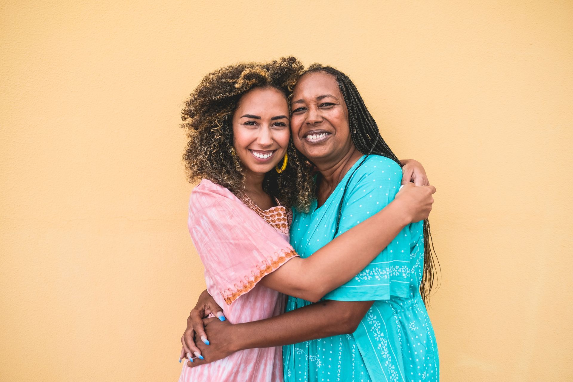 Woman hugs her mother in front of a yellow wall; both smile. The woman wears pink, and her mother wears blue.