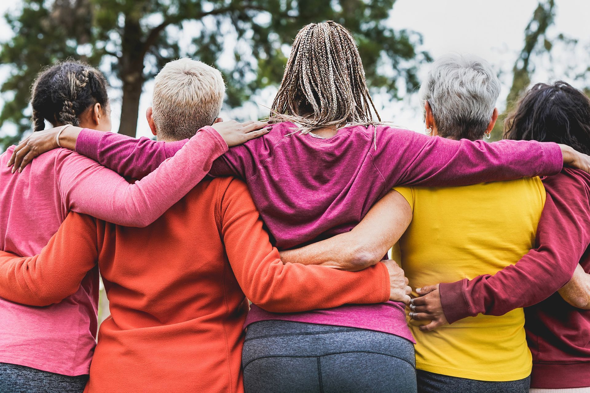 Five women with arms around each other, wearing colorful shirts outdoors.