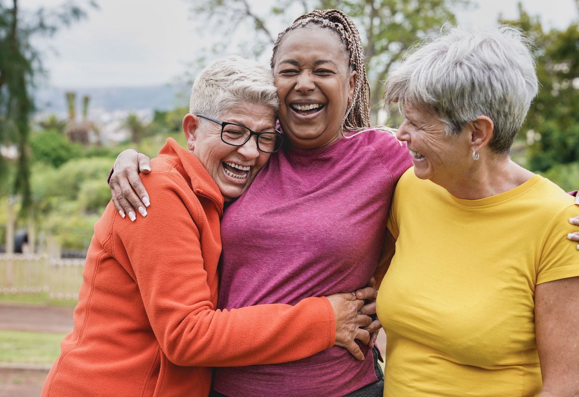 Three women of color laughing and hugging outside; one in orange, one in pink, and one in yellow.