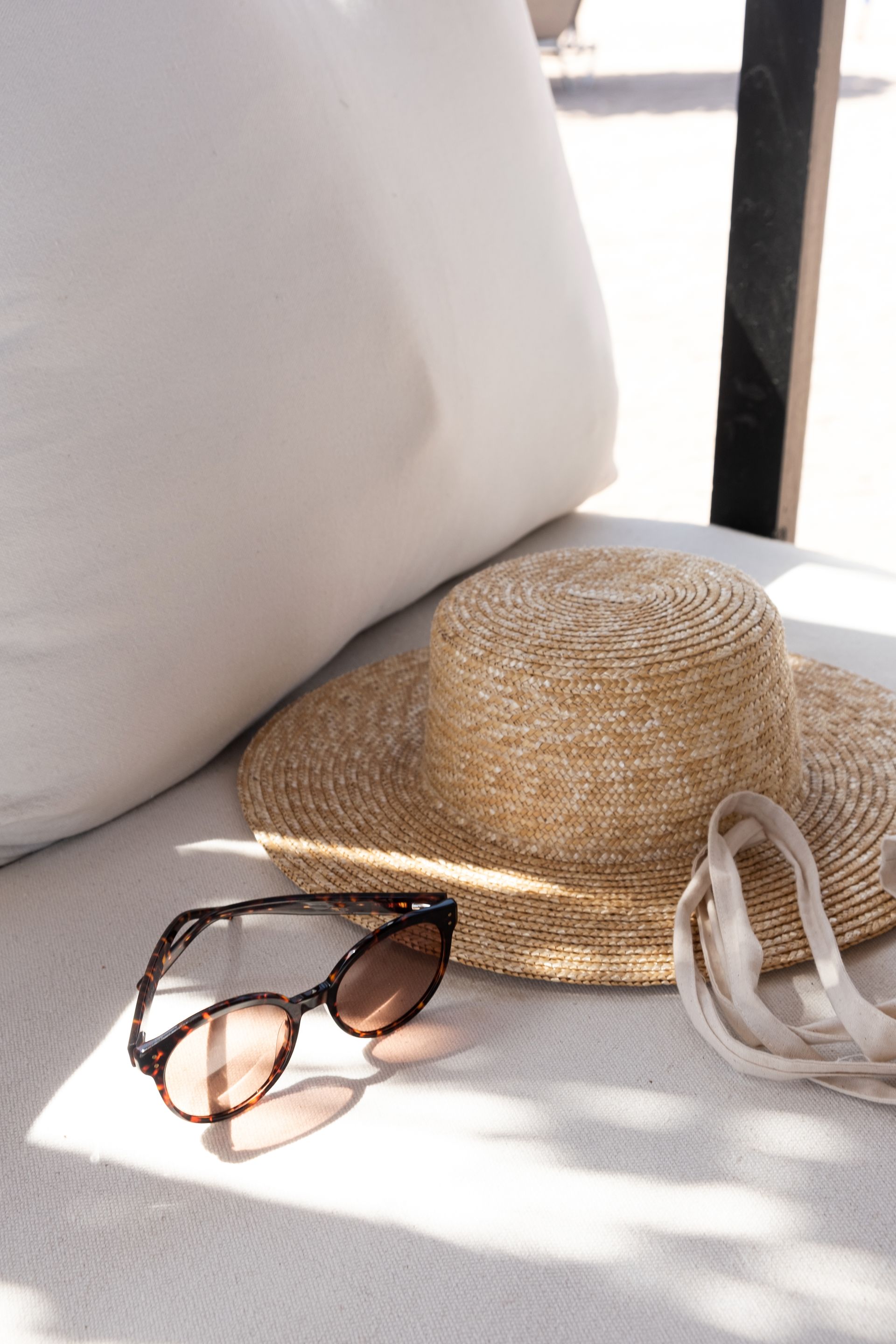 Straw hat, sunglasses, and bag resting on a white cushion in sunlight.