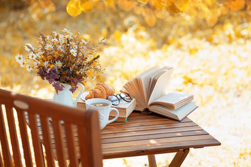 Outdoor table set for a cozy break: flowers, croissant, tea, books, and glasses in warm autumn light.