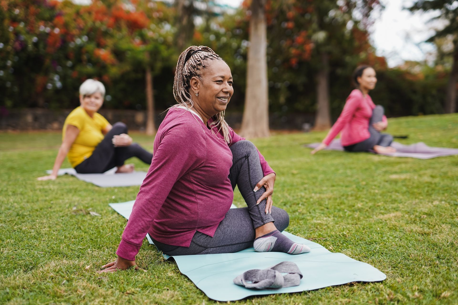 Three women practicing yoga on mats in a grassy park; one twists body, smiling.